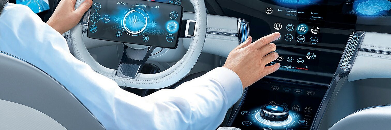 A person in a white jacket operates a futuristic car with digital displays and touch controls. The dashboard shows various electronic interfaces. The view outside suggests a cityscape with modern buildings. All exposed wiring within the vehicle has been neatly secured using tesa tape, ensuring a clean and organized appearance.