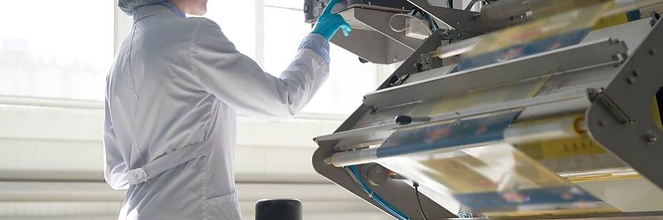 A person in a white lab coat and hair net operates a packaging machine. They are pressing a button or touch screen on the device. The machine is processing a roll of transparent tesa tape. Sunlight streams through windows in the background.