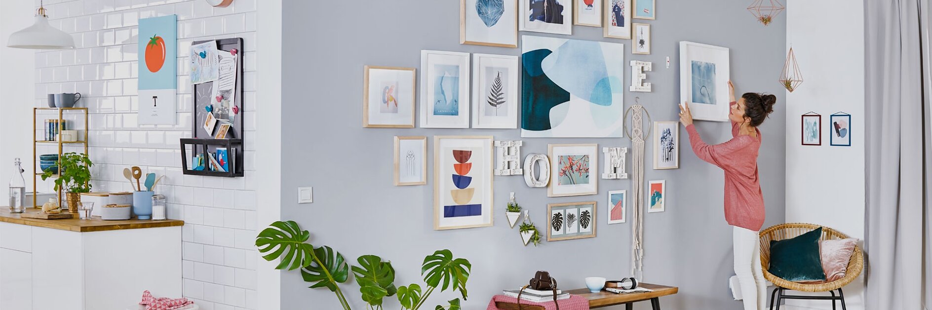A woman in a pink shirt is arranging frames on a grey and white wall, filled with various artwork and photos, using tesa tape. The room has a modern design with a small kitchen on the left, light wood flooring, and green plants for decoration.