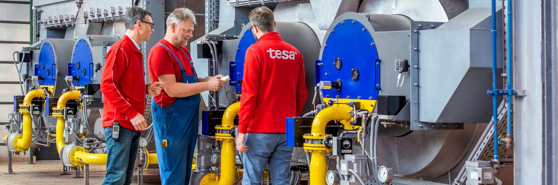 Three men stand in front of large industrial machinery with blue and silver components. They are wearing red shirts and are having a discussion. Yellow pipes are connected to the machines, and the floor is paved with tiles.