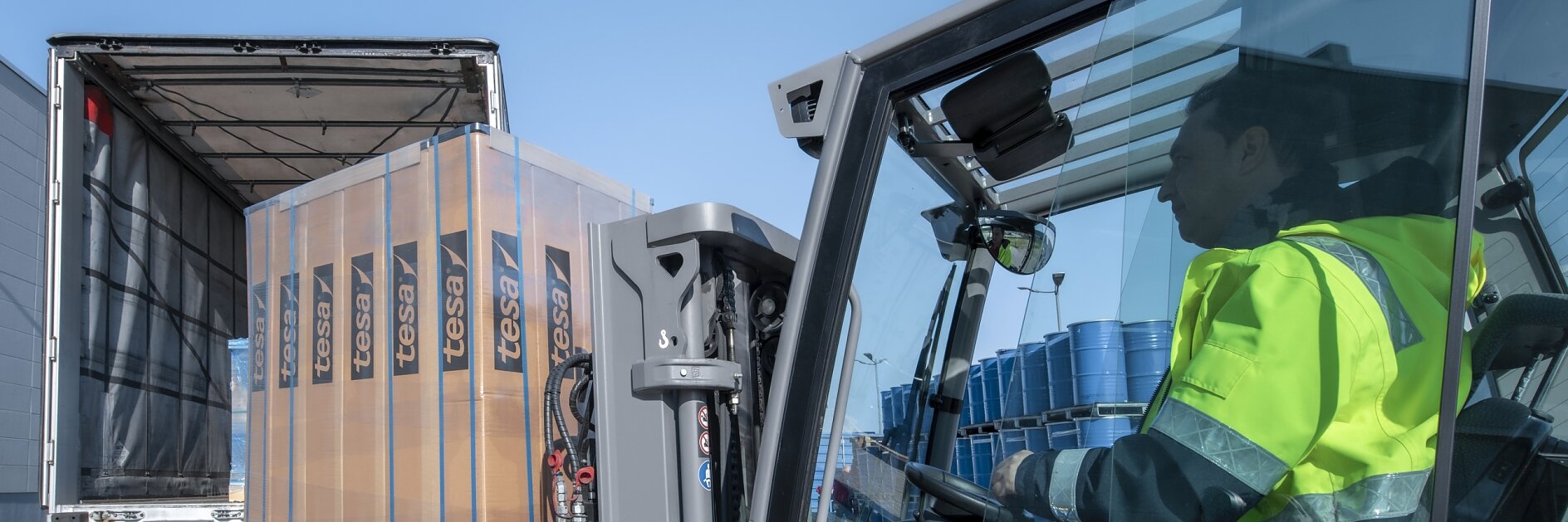 A forklift operator wearing a fluorescent yellow jacket loads a pallet of wrapped goods labeled Fessi into the back of a partially open truck. Blue barrels are visible in the truck. The scene takes place outdoors on a clear day, and everything is securely fastened with tesa tape.