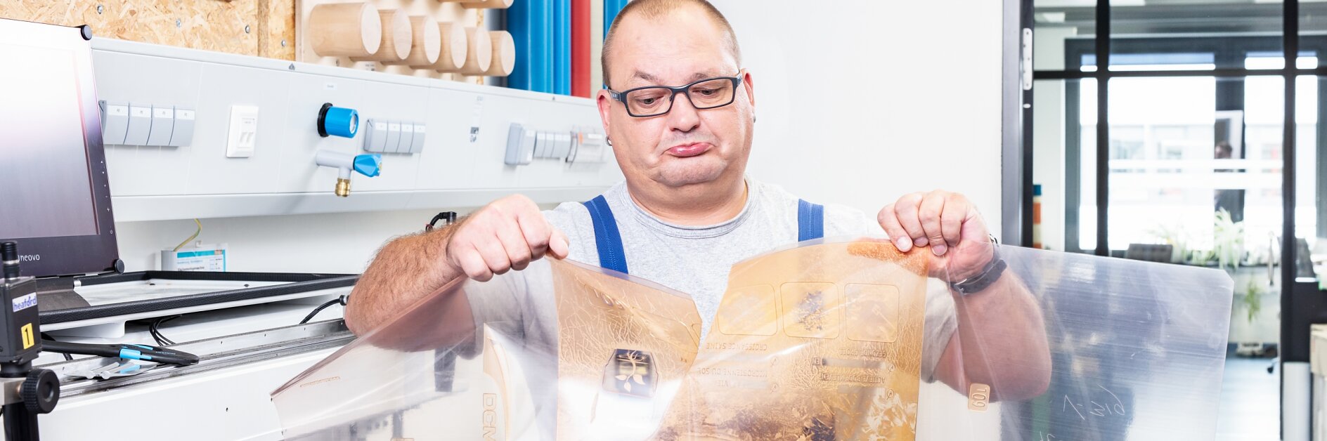 A man wearing glasses and a gray shirt is holding a large, transparent plastic sheet with printed designs secured using tesa tape. He stands in a workroom with machinery, wooden cylinders on shelves, and a visible doorway leading to another area.