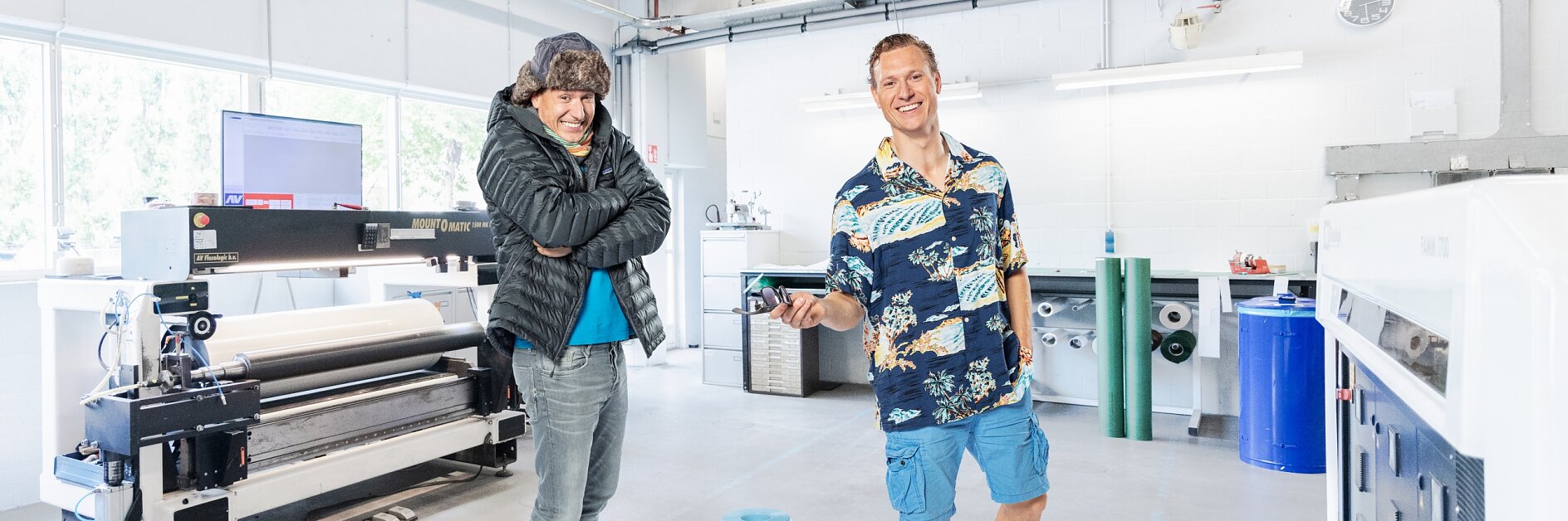 Two people stand in a bright workspace. One wears a beanie and puffy jacket, crossing their arms, while the other sports a Hawaiian shirt and shorts. A large printer is in the background along with some rolled materials. One of them looks serious, while the other is smiling.