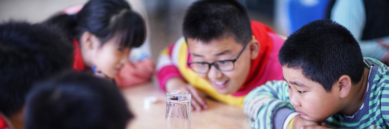 Children are gathered around a table, observing a science experiment. A lit candle is on a plate covered by an inverted glass, secured in place with tesa tape. One child is pointing at it, while the others watch attentively, suggesting they are curious about the experiment.