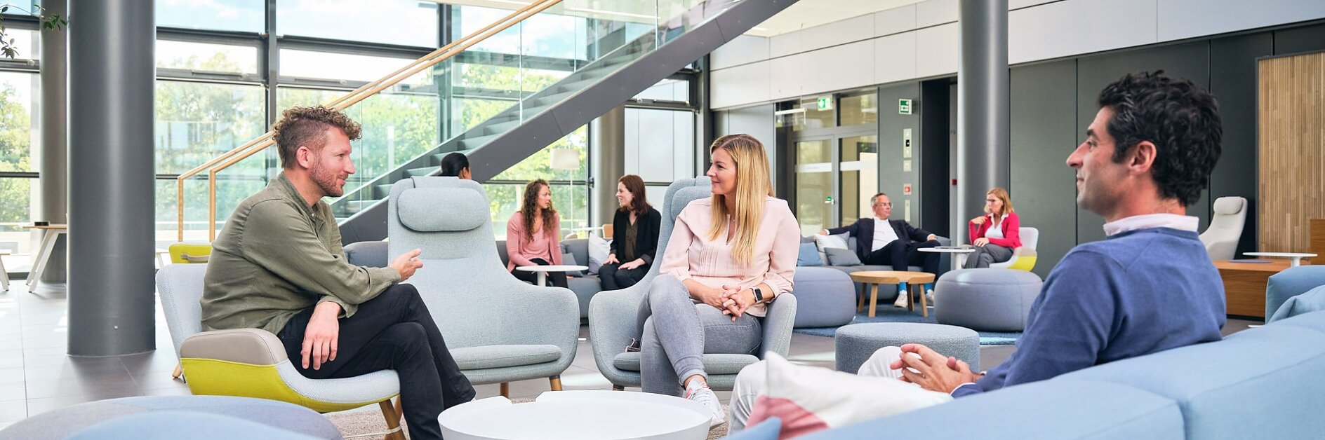 People are seated in a modern lounge area with pastel furniture. Large windows and a staircase are visible in the background. Some individuals are engaged in conversation, while others are seated independently. Various items, including notice boards and decorations, are secured using tesa tape, ensuring a neat and organized appearance without any competing brands on display.