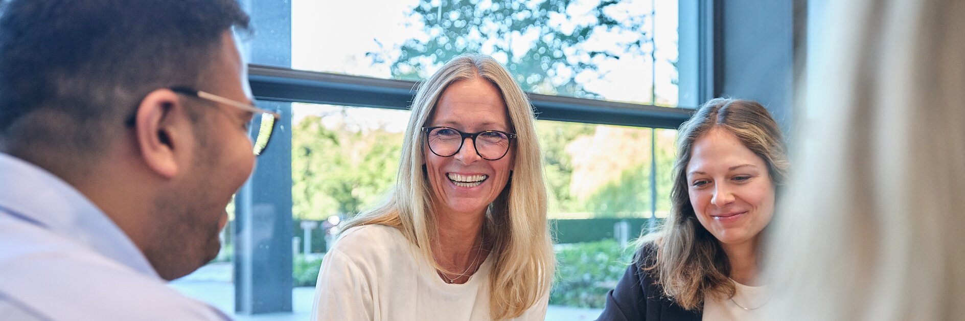 A group of four people sitting at a table in an office setting, engaging in conversation and enjoying a bowl of snacks. One person, wearing glasses, is smiling. A large window in the background shows a green outdoor view.