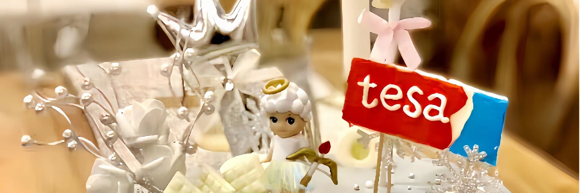 A cake with white frosting topped with a small angel figurine, a red label with the word tesa, white flowers, and decorative silver elements. A silver balloon and crown are in the background.