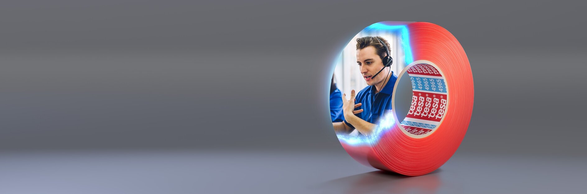 A red roll of tesa tape is on a gray surface. The side of the tesa tape features an image of a person wearing a headset, gesturing with one hand. Next to the person is a blue and red calendar with some dates marked.