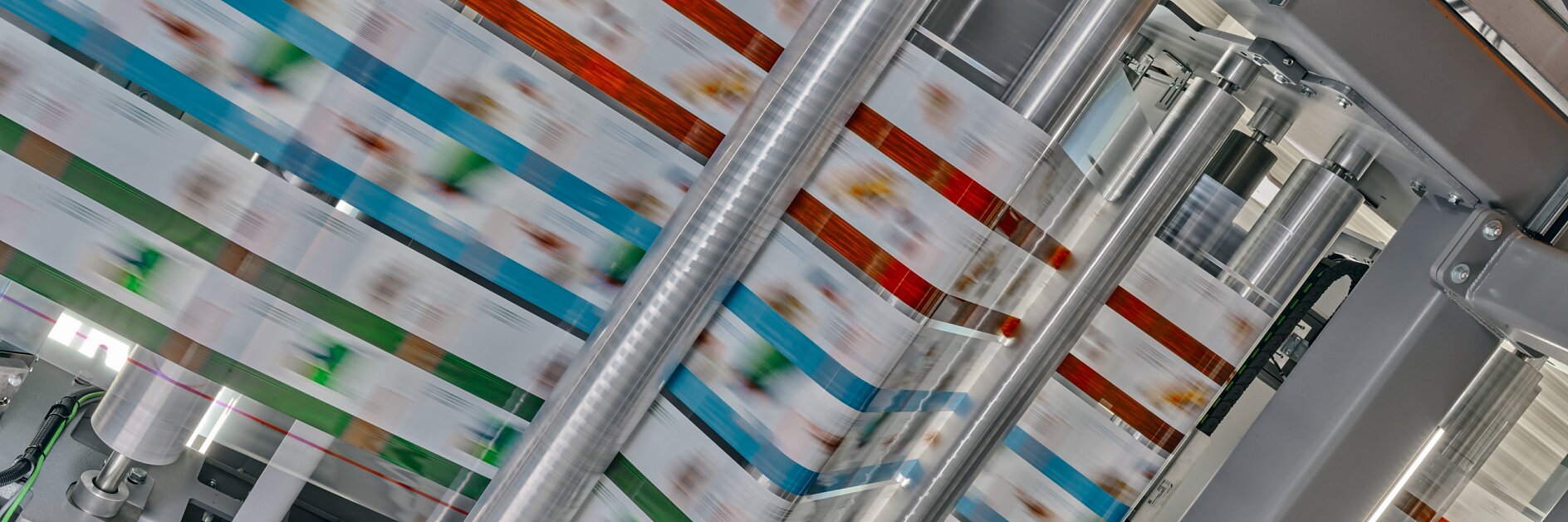A high-speed printing press produces colorful packaging material. Multiple rollers guide the printed material through the machine, showing a blur of colors, including red, blue, and green patterns, under bright lighting. The process relies on the use of tesa tape to ensure everything stays in place during production.