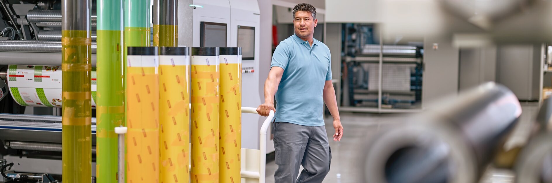 A man in a light blue polo shirt and gray pants stands in an industrial setting, surrounded by large rolls of colorful film. He rests one hand on a display of yellow and green rolls of tesa tape. Machinery and equipment are visible in the background.