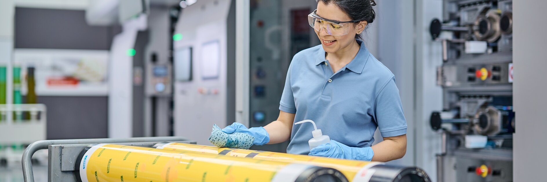 A person wearing safety glasses and gloves stands beside a printing press machine, cleaning large yellow rollers with a cloth and spray bottle. The scene appears to be in an industrial or manufacturing setting, with tesa tape nearby for additional tasks.