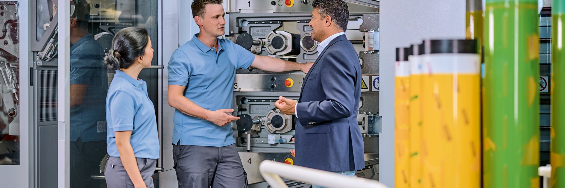 Three people stand in front of industrial machinery. Two individuals wear blue work uniforms, and one person in a blue blazer and jeans gestures while speaking. Rolls of tesa tape are visible in the foreground.