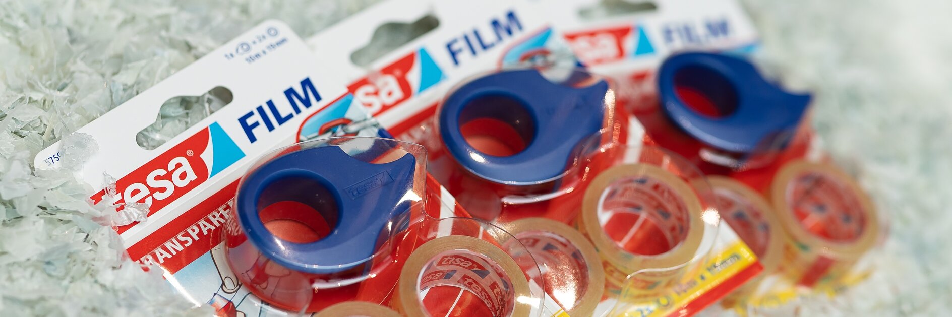 Three packages of transparent tesa tape with blue dispensers are displayed. Each package includes three rolls of tesa tape and features brand labeling. The background is covered with shredded white material.