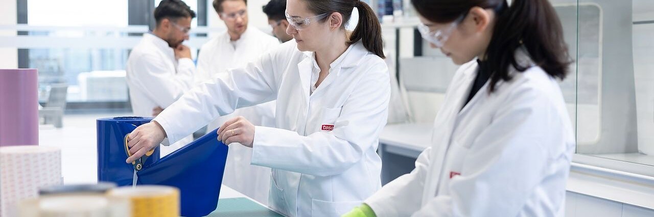 Several people in lab coats and safety glasses work with large rolls of material and tesa tape on tables in a bright laboratory, with shelves and lab equipment visible in the background.