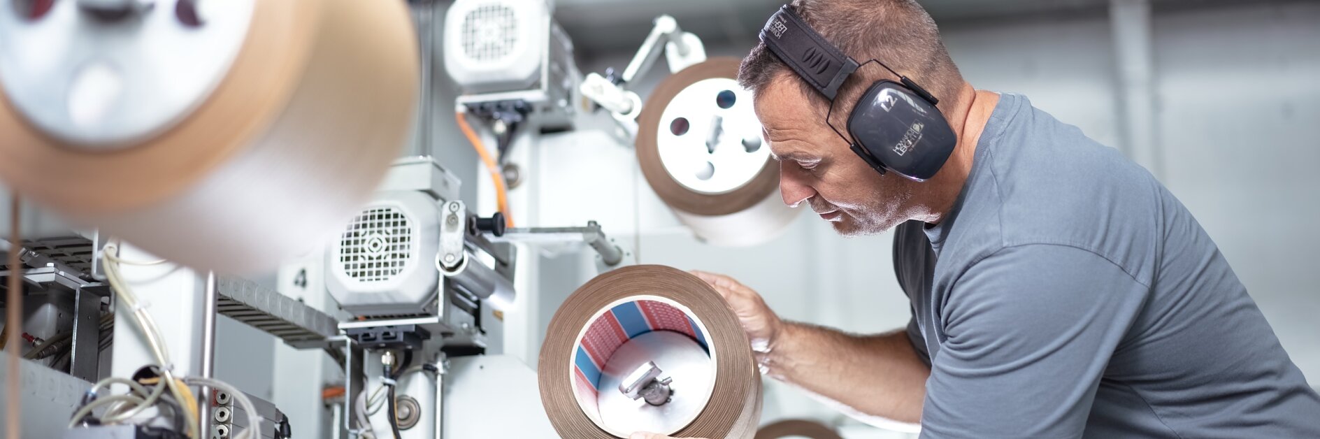 A man wearing ear protection inspects a roll of material in an industrial setting. Various machines with similar rolls of tesa tape are visible in the background. He is focused on examining the roll closely.