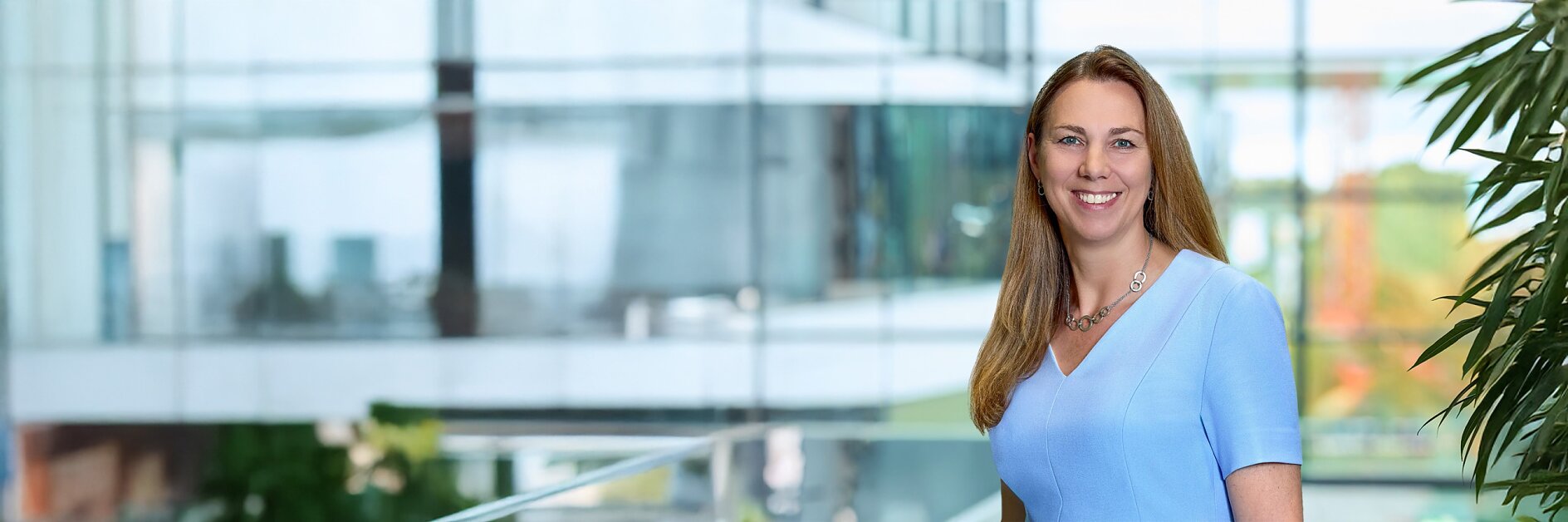 A woman with long brown hair, wearing a light blue short-sleeve top and a necklace, stands indoors by glass windows and green plants, smiling.